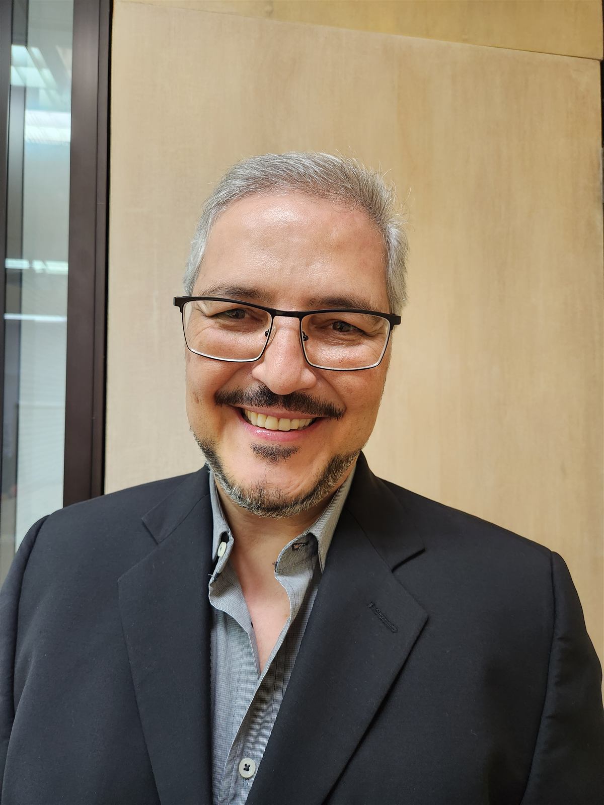 Retrato de Ricardo Lopes sorrindo, de terno, ao lado de uma planta no consultório.